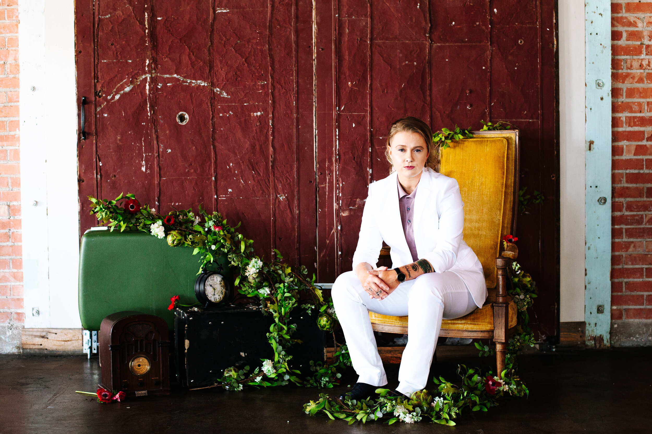  Bride in white suit sits in a vintage gold-yellow chair surrounded by greenery at her lesbian wedding.