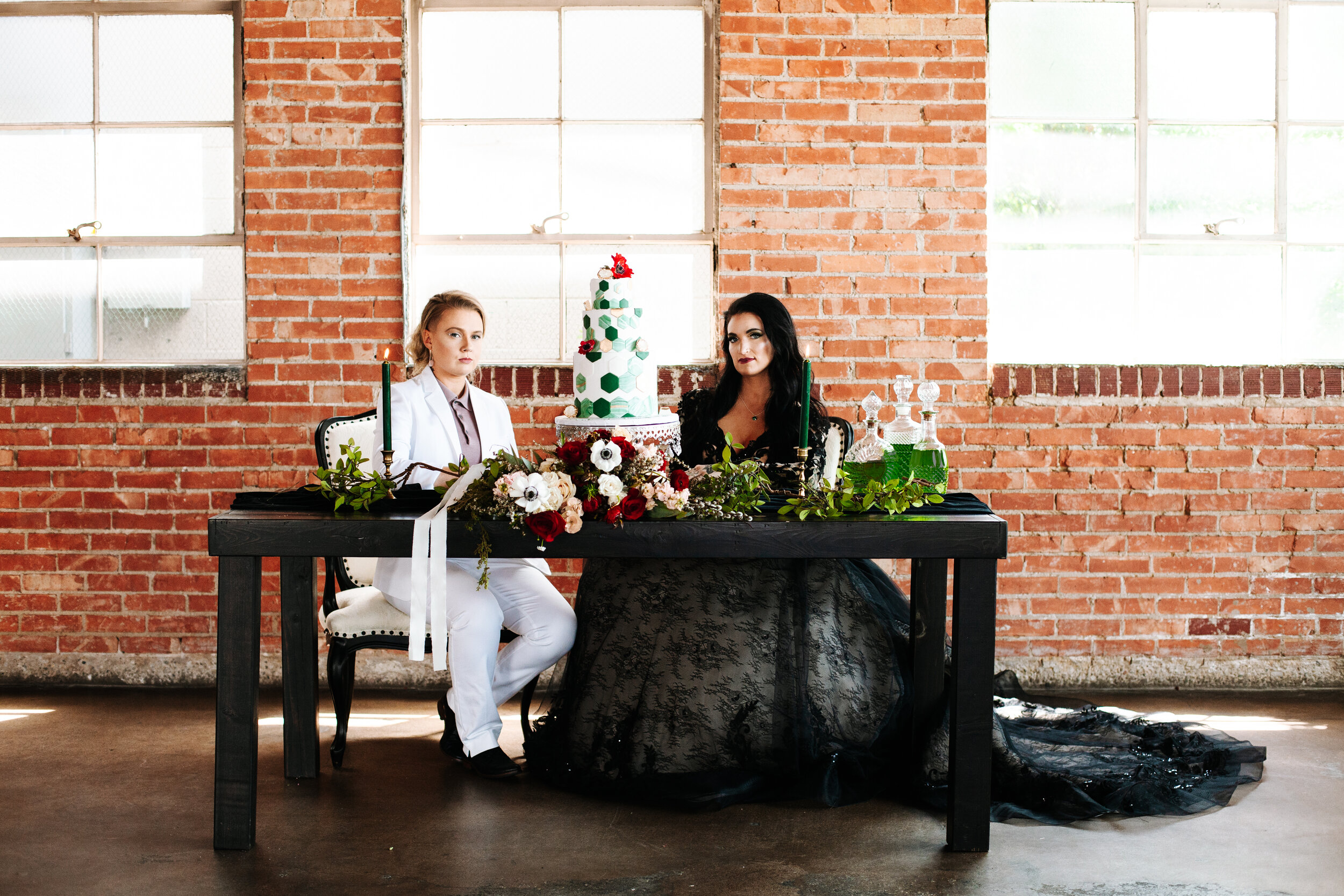 Lesbian couple sit together for their wedding at a sweetheart table decorated with greenery and a bouquet. 