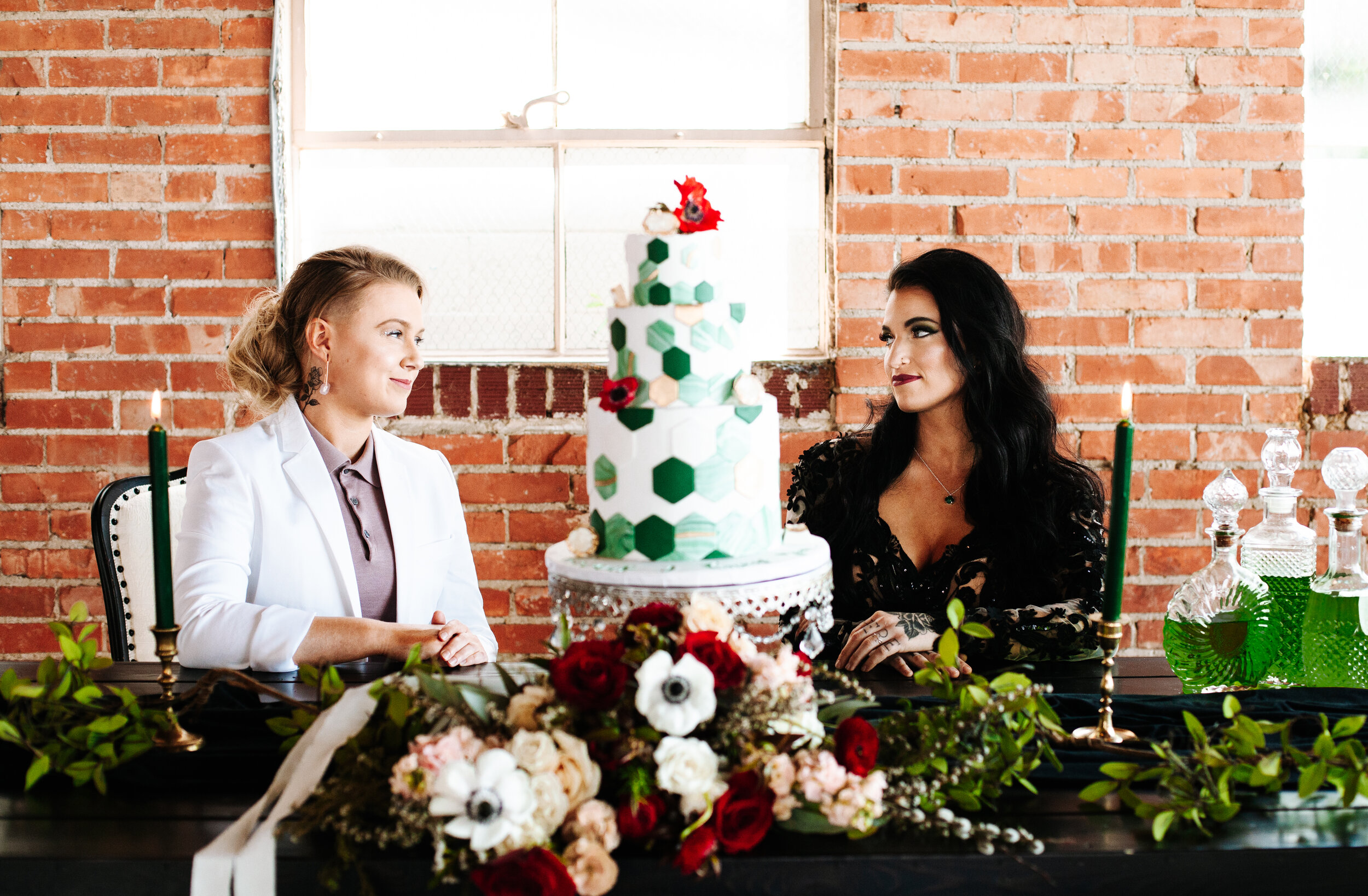 Lesbian couple looking at each other at their wedding. They're sitting at a sweetheart table decorated with greenery and a bouquet. 