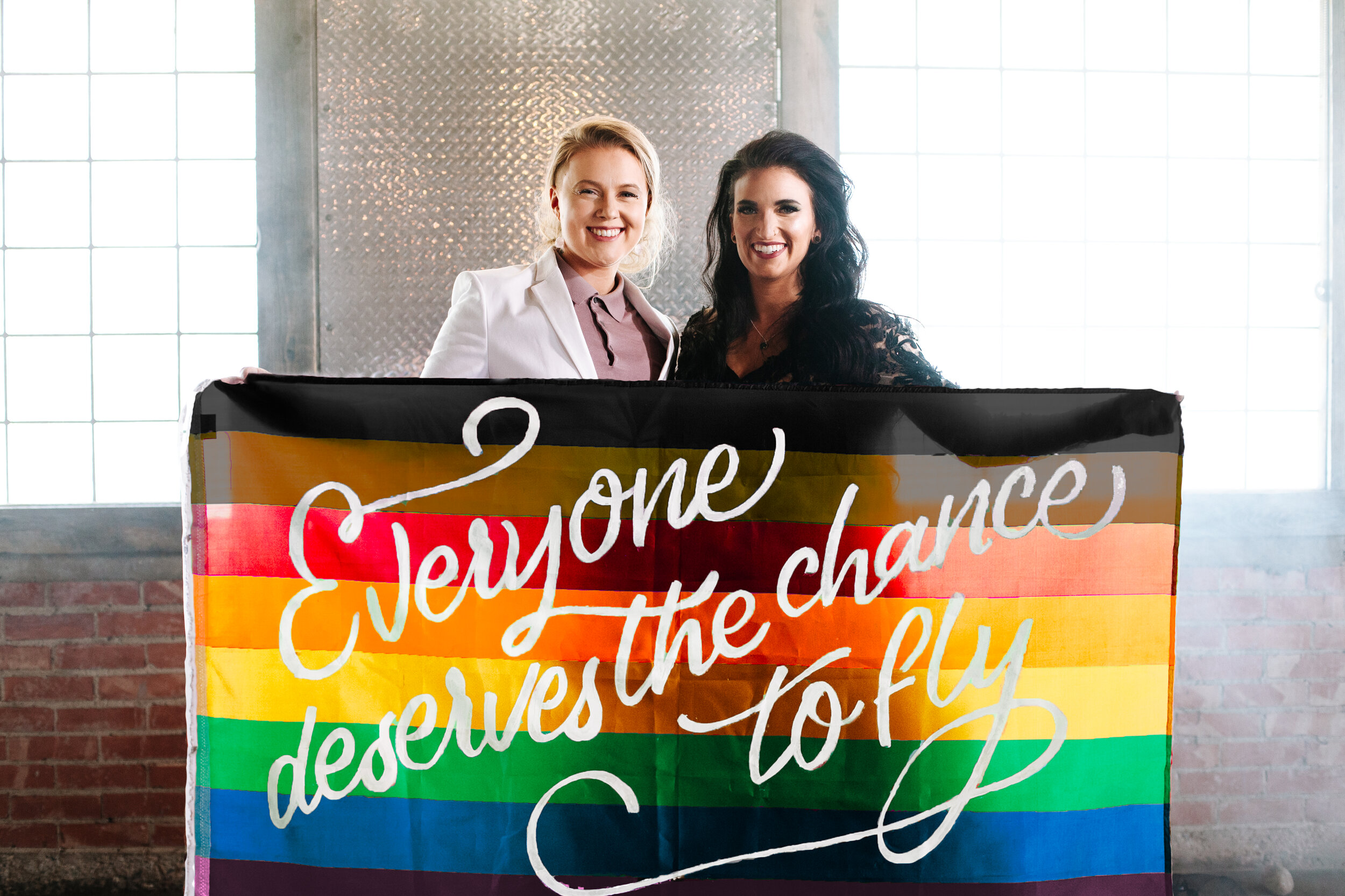 Lesbian couple looking at each other at their wedding holding a pride flag that says, "Everyone deserves the chance to fly"-a quote from the broadway play Wicked.
